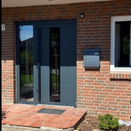 PS OKNA Przemysław Szustek - Nowoczesne, grafitowe drzwi wejściowe z przeszkleniami, zamontowane w ceglanym budynku z numerem 6. Obok skrzynka pocztowa w tym samym kolorze. Widoczne płytki wejściowe.