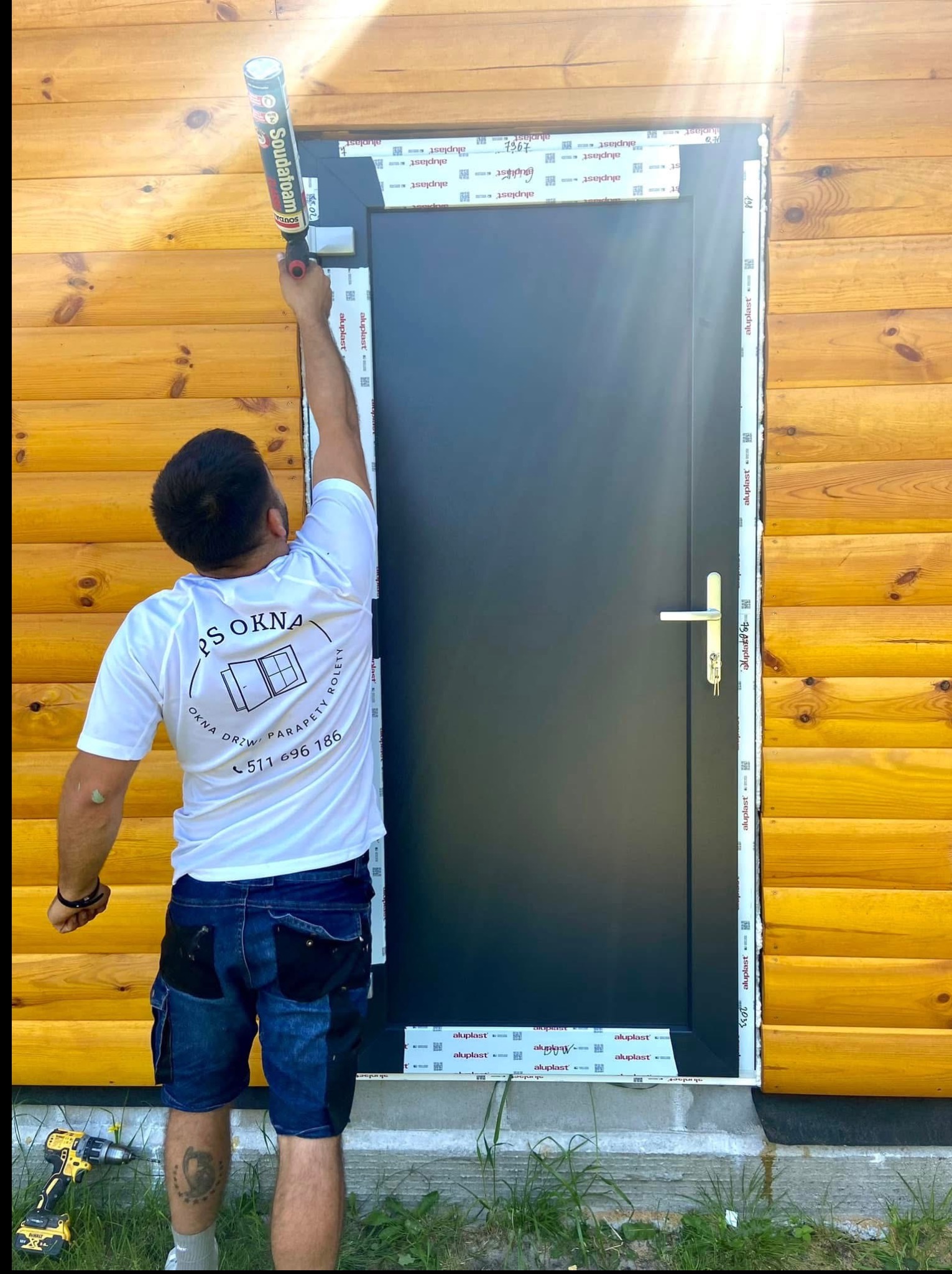 Montaż drzwi zewnętrznych: monter uszczelnia pianką ramę czarnych drzwi na tle drewnianej elewacji. Widoczny profil Aluplast i narzędzia pracy.