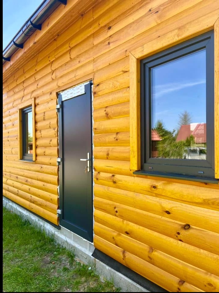 Dom z bali z czarnymi drzwiami i oknami PCV z widocznym odbiciem nieba. Elewacja w ciepłym odcieniu drewna, nowoczesny design, estetyczne wykończenie.