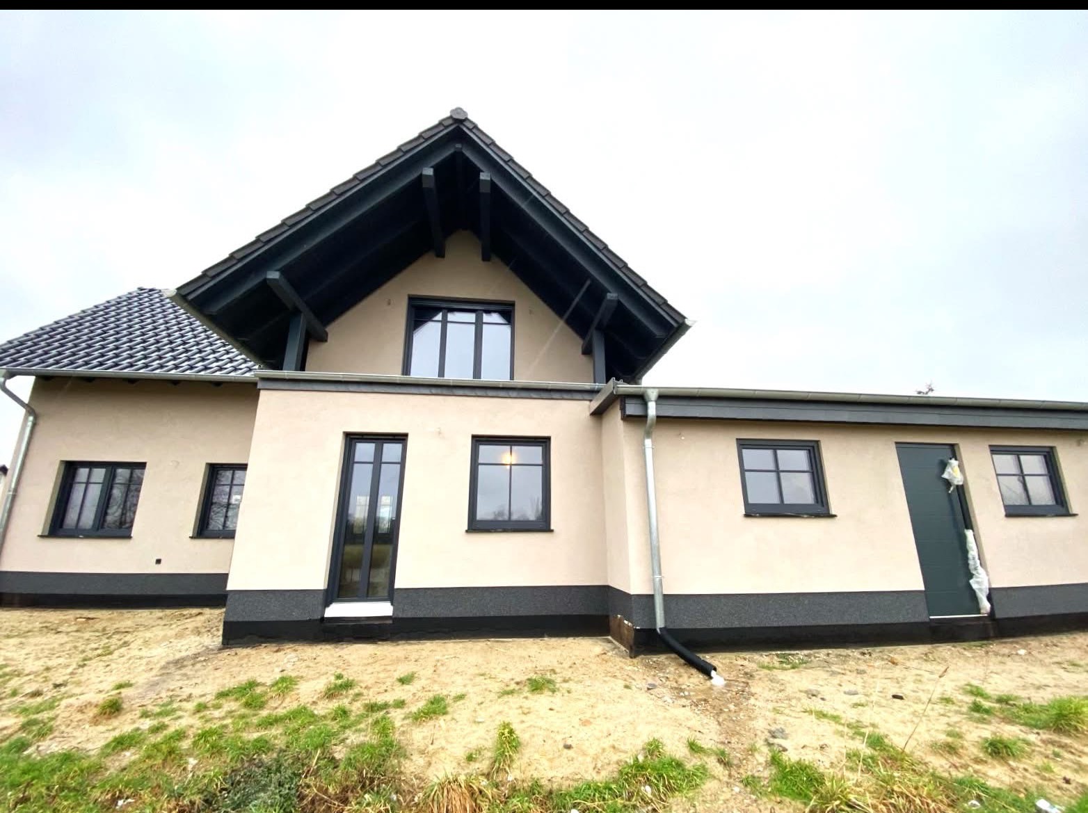 Nowoczesny dom jednorodzinny z ciemnymi oknami i drzwiami na tle pochmurnego nieba. Widoczne orynnowanie i fragment terenu z zielenią. Minimalistyczny design.