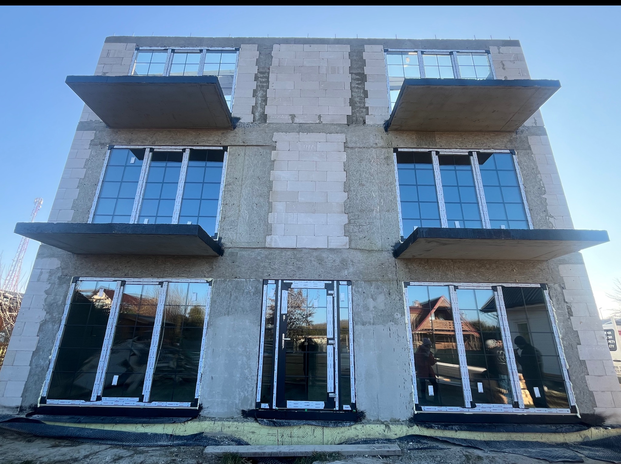 Surowy budynek z zamontowanymi oknami z białymi ramami i betonowymi balkonami, w trakcie budowy, widok z dołu na błękitne niebo.