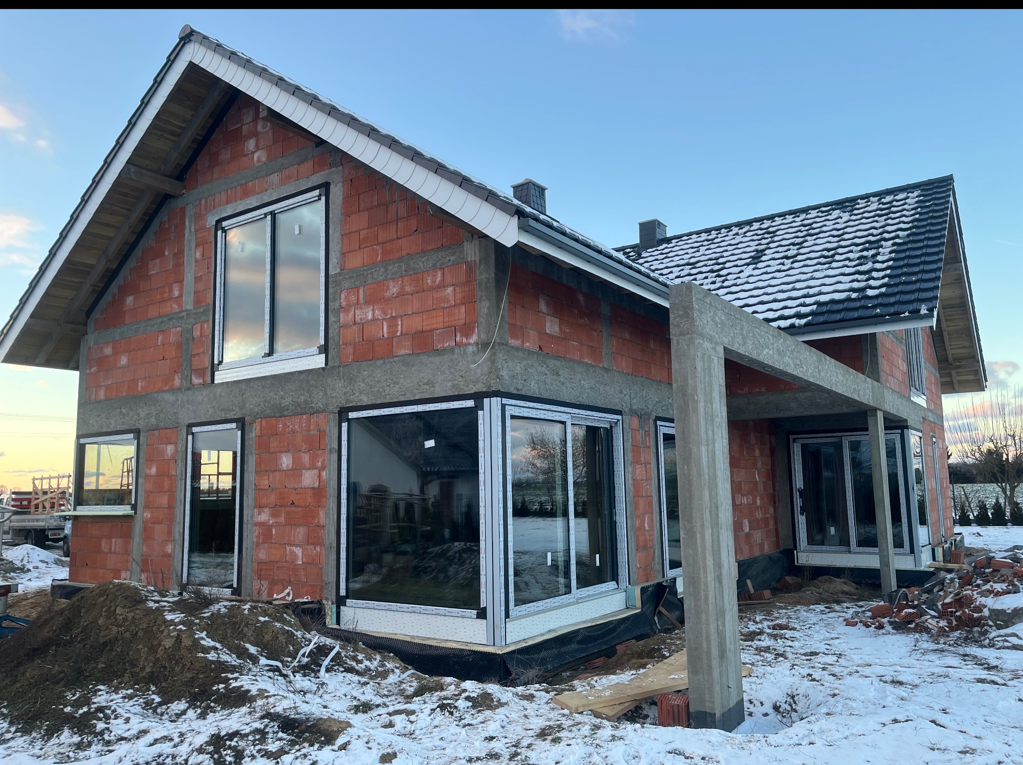 Nowy dom w trakcie budowy z zamontowanymi oknami PCV. Widoczne cegły, betonowe nadproża, zasypany śniegiem teren. Okna z szybami odbijają niebo.