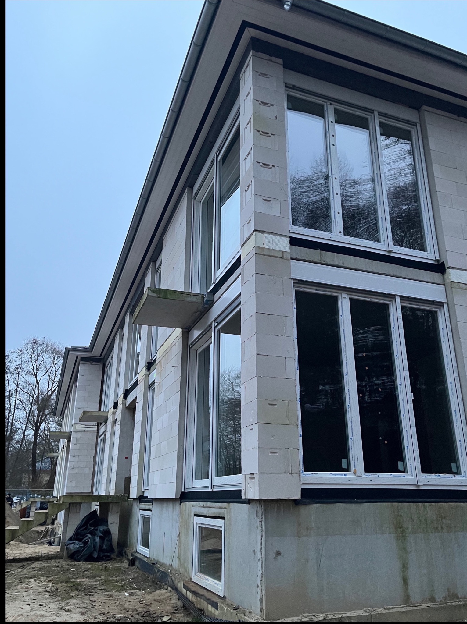 Surowy budynek z białego betonu komórkowego z dużymi oknami PCV, widok z dołu na budynek w trakcie budowy, z widocznym ociepleniem fundamentów.