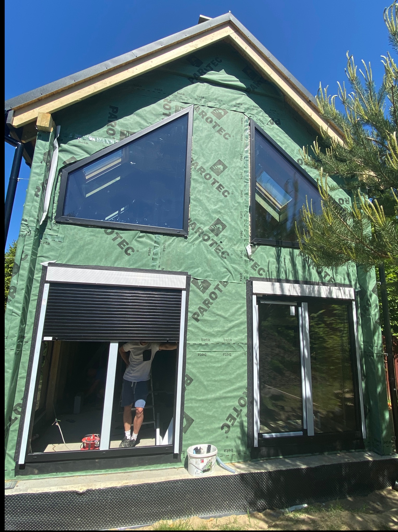 Budynek w trakcie montażu stolarki okiennej z roletami zewnętrznymi, widoczne okna o nietypowych kształtach i folia paroizolacyjna na ścianach.