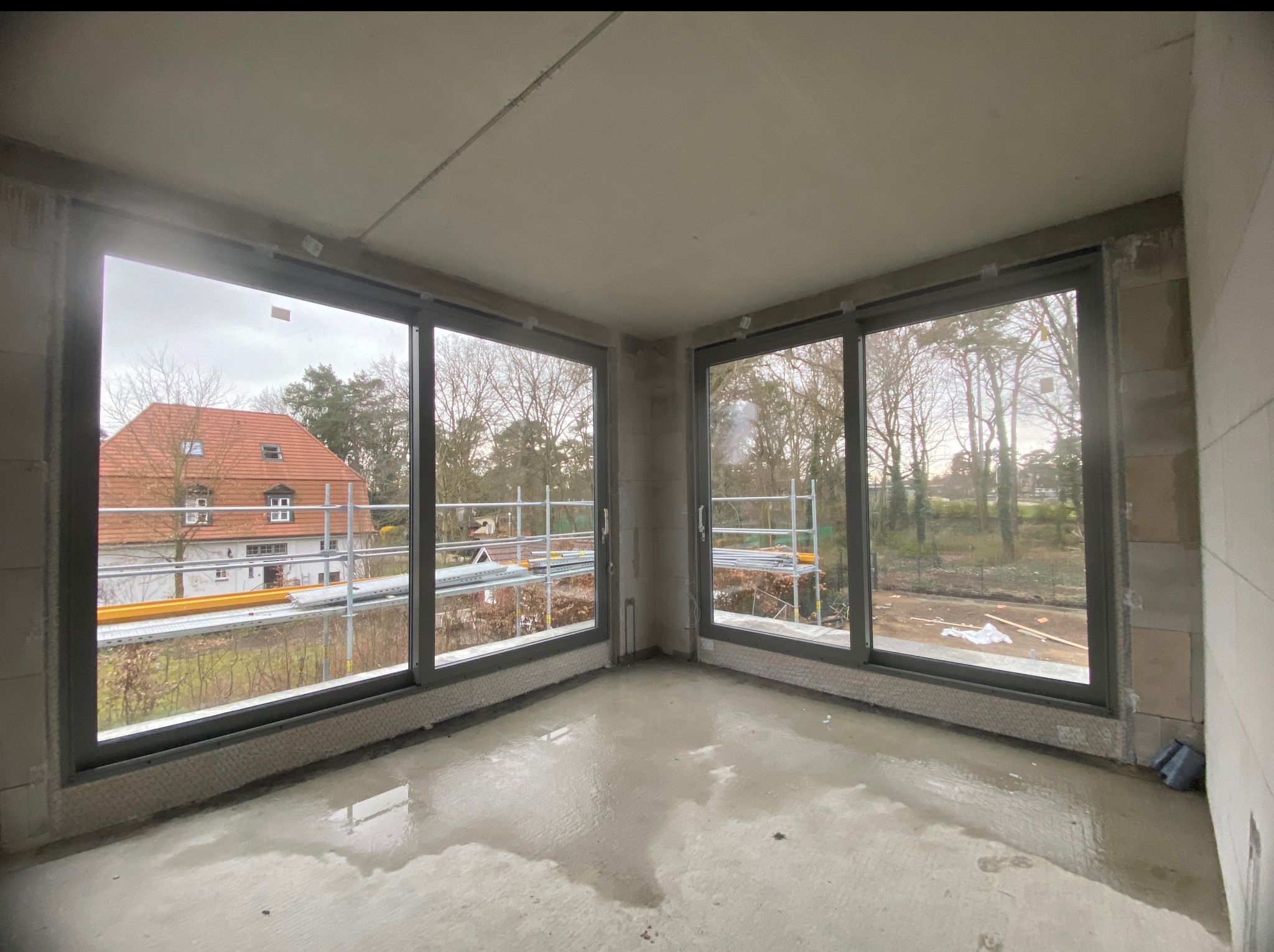 Wnętrze surowego pomieszczenia z dwoma oknami PCV z widokiem na rusztowanie i budynek z czerwoną dachówką. Widoczne bloczki betonowe i mokra posadzka.