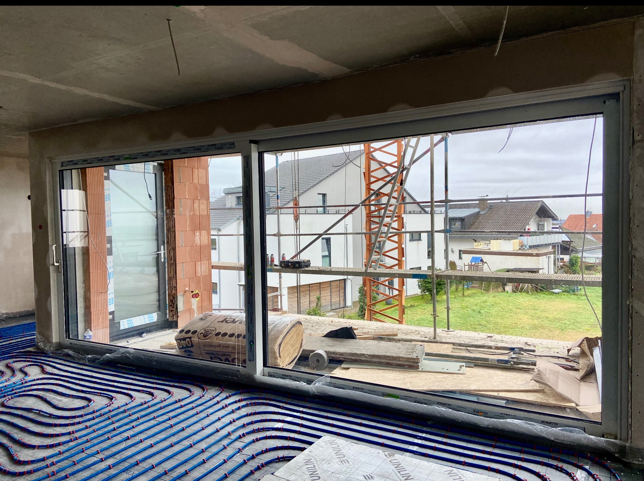 Wnętrze w trakcie budowy z widokiem na zewnątrz przez duże okno przesuwne. Widoczne ogrzewanie podłogowe, cegły i materiały budowlane. Na zewnątrz rusztowanie i domy.