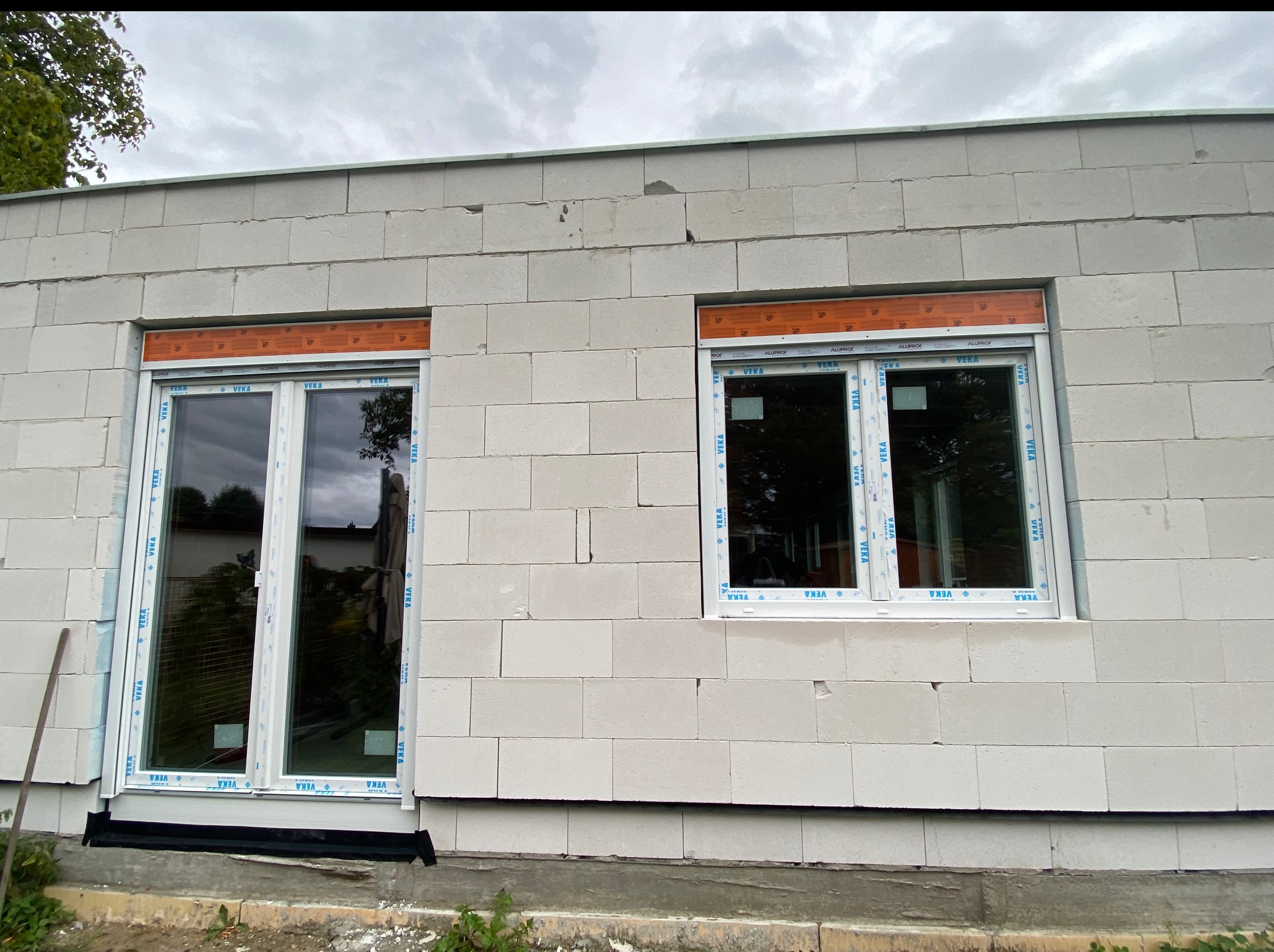 Montaż okien PCV w surowym budynku z betonu komórkowego. Widoczne zabezpieczenia na ramach okiennych. Okno i drzwi balkonowe zamontowane obok siebie.