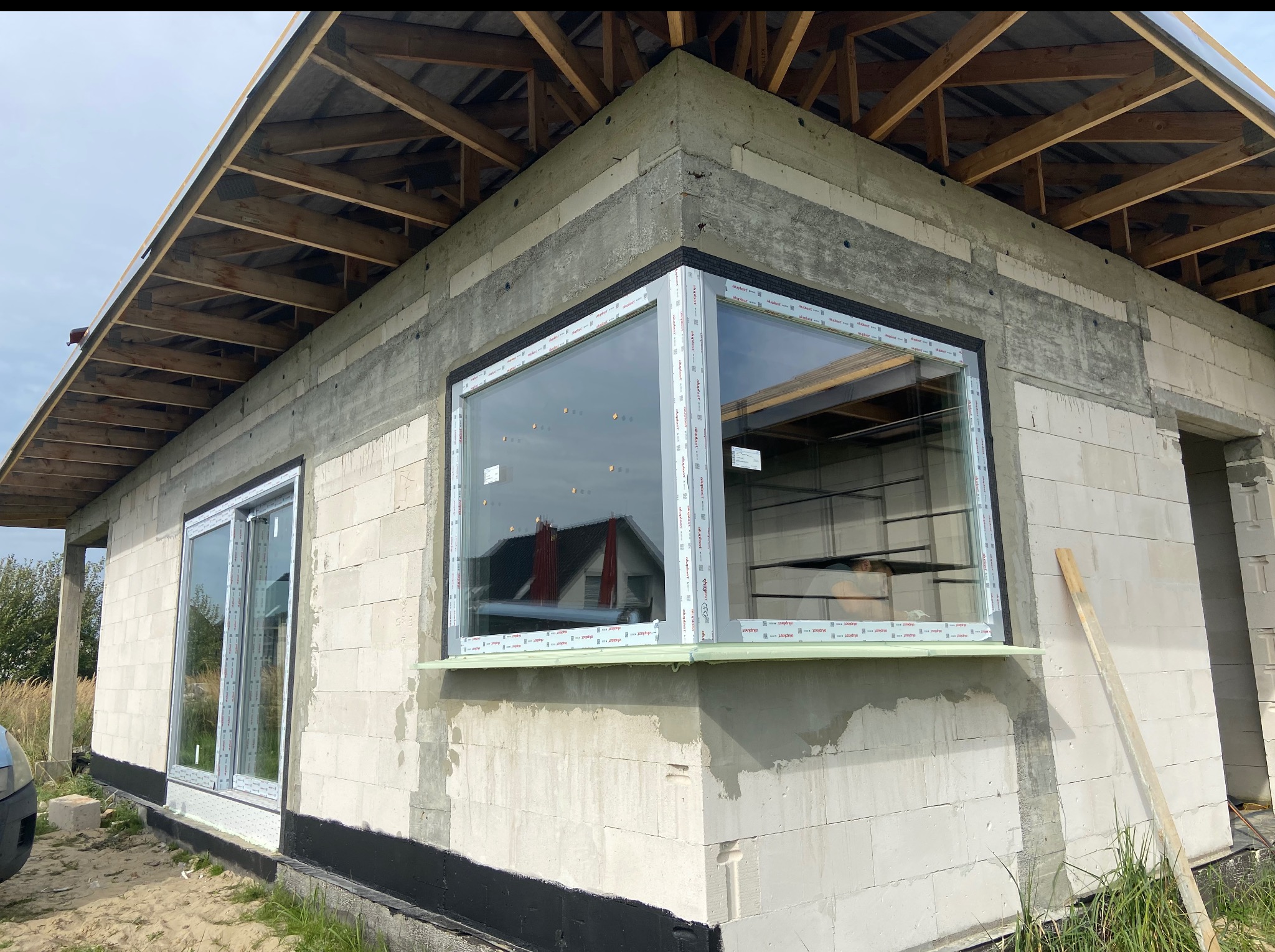 Surowy budynek z zamontowanymi oknami PCV: narożne okno z szeroką ramą i podwójne okno obok. Widoczna konstrukcja dachu. W tle dom z czerwonym dachem.