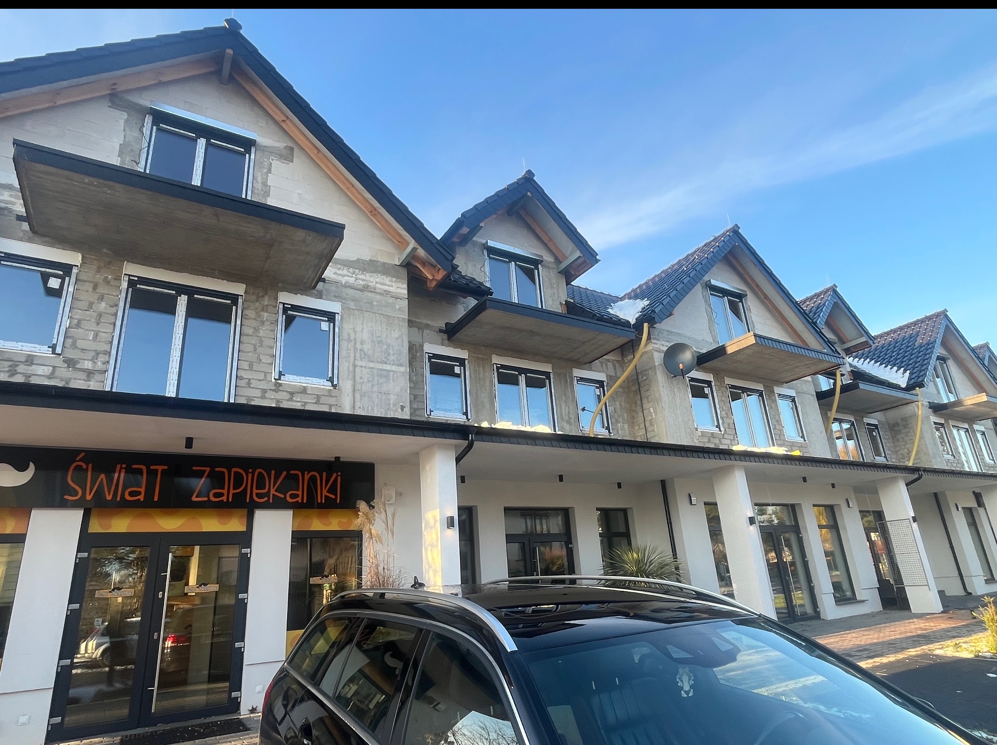 Budynek w trakcie wykończenia z zamontowanymi oknami, widoczne balkony i fragment zaparkowanego samochodu na pierwszym planie, błękitne niebo w tle.