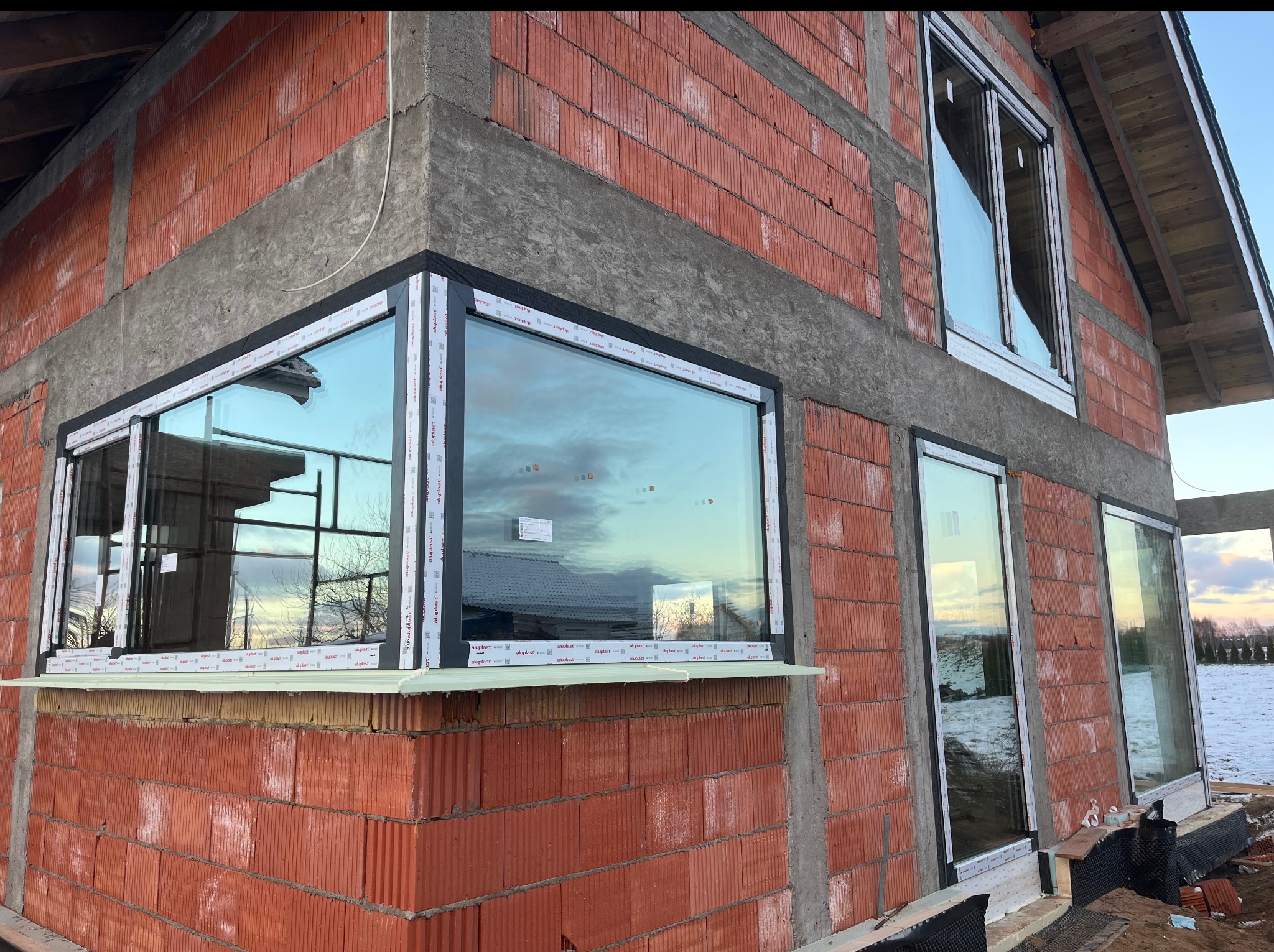 Surowy stan budynku z czerwonej cegły z zamontowanymi oknami PCV marki Aluplast. Widoczne parapety i wstępne wykończenie elewacji. Częściowo ośnieżone otoczenie.