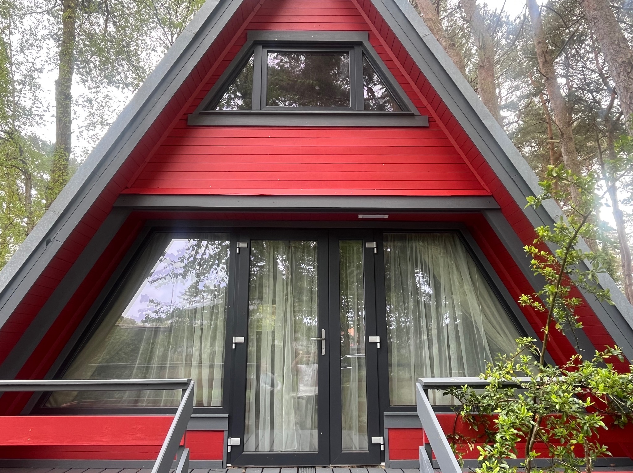 Czerwony domek w stylu A-frame z szarymi detalami, widoczne duże okna i drzwi balkonowe z firanami, otoczony zielenią.