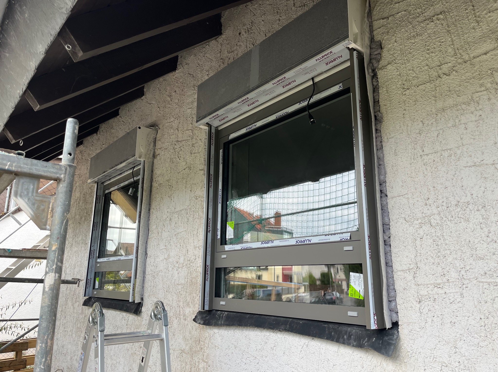 Montaż okien z roletami zewnętrznymi w szarej ramie. Widoczna piana montażowa uszczelniająca okna. Na pierwszym planie drabina i rusztowanie.