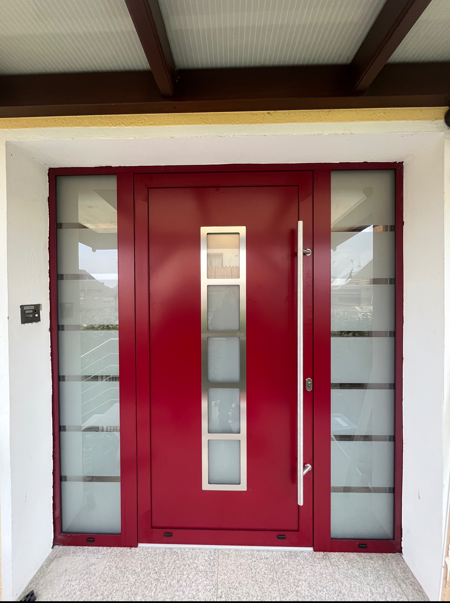 Czerwone drzwi wejściowe z szybami z poziomymi paskami, długa srebrna klamka, nowoczesny design.