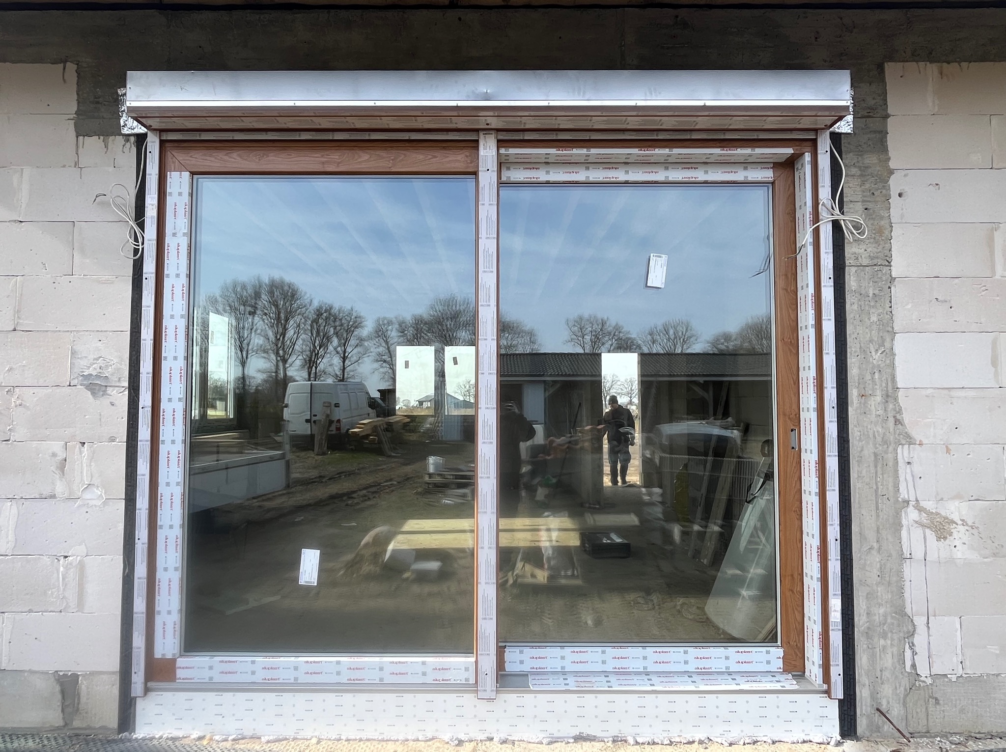 Brązowe okno z roletą zewnętrzną zamontowane w surowym murze z betonu komórkowego. Widoczne elementy montażowe i folia ochronna.