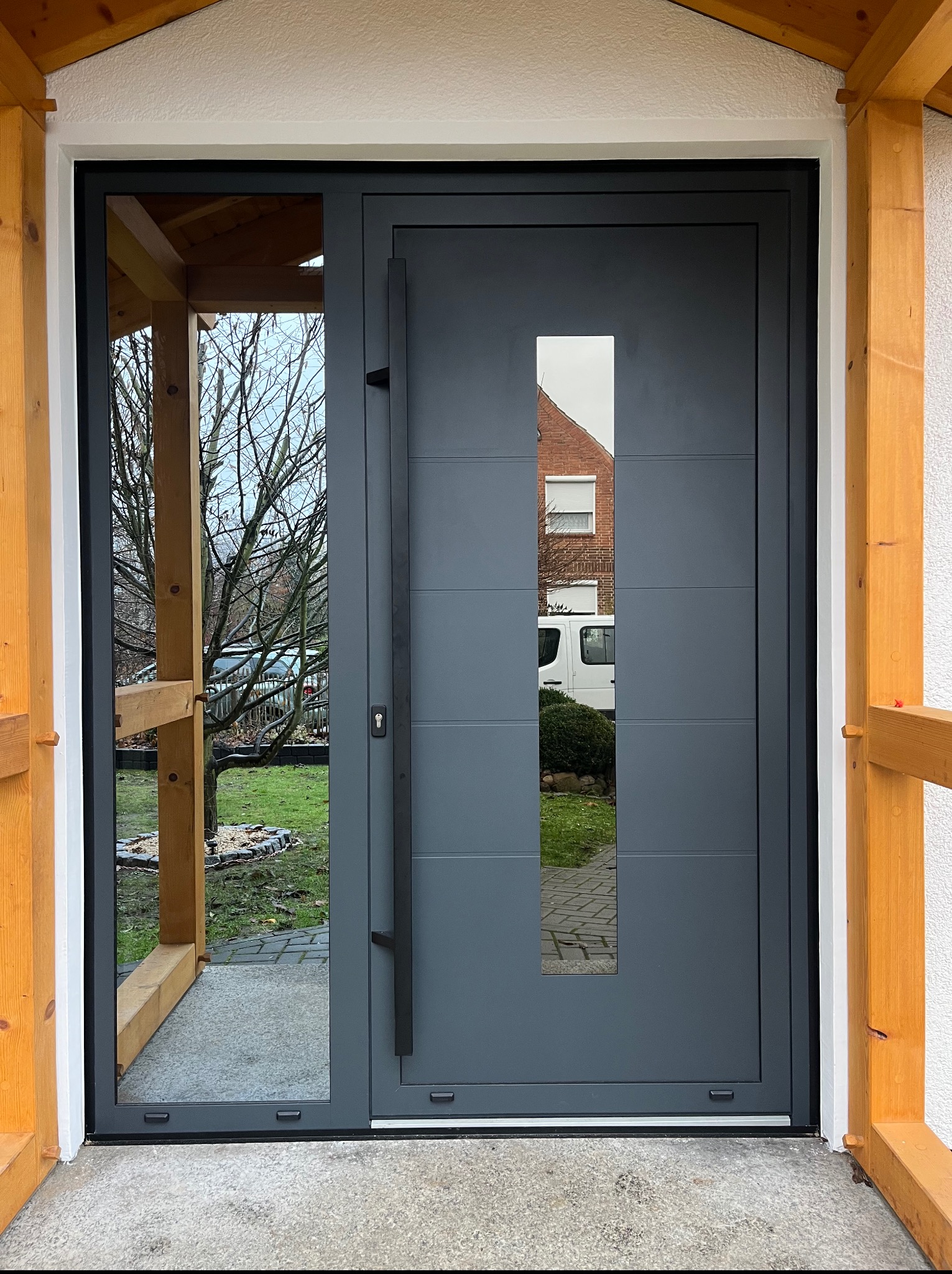 Szare drzwi zewnętrzne z długim uchwytem i szybą, widok z zewnątrz, pod zadaszeniem z drewnianą konstrukcją. W szybie odbija się budynek z roletami.