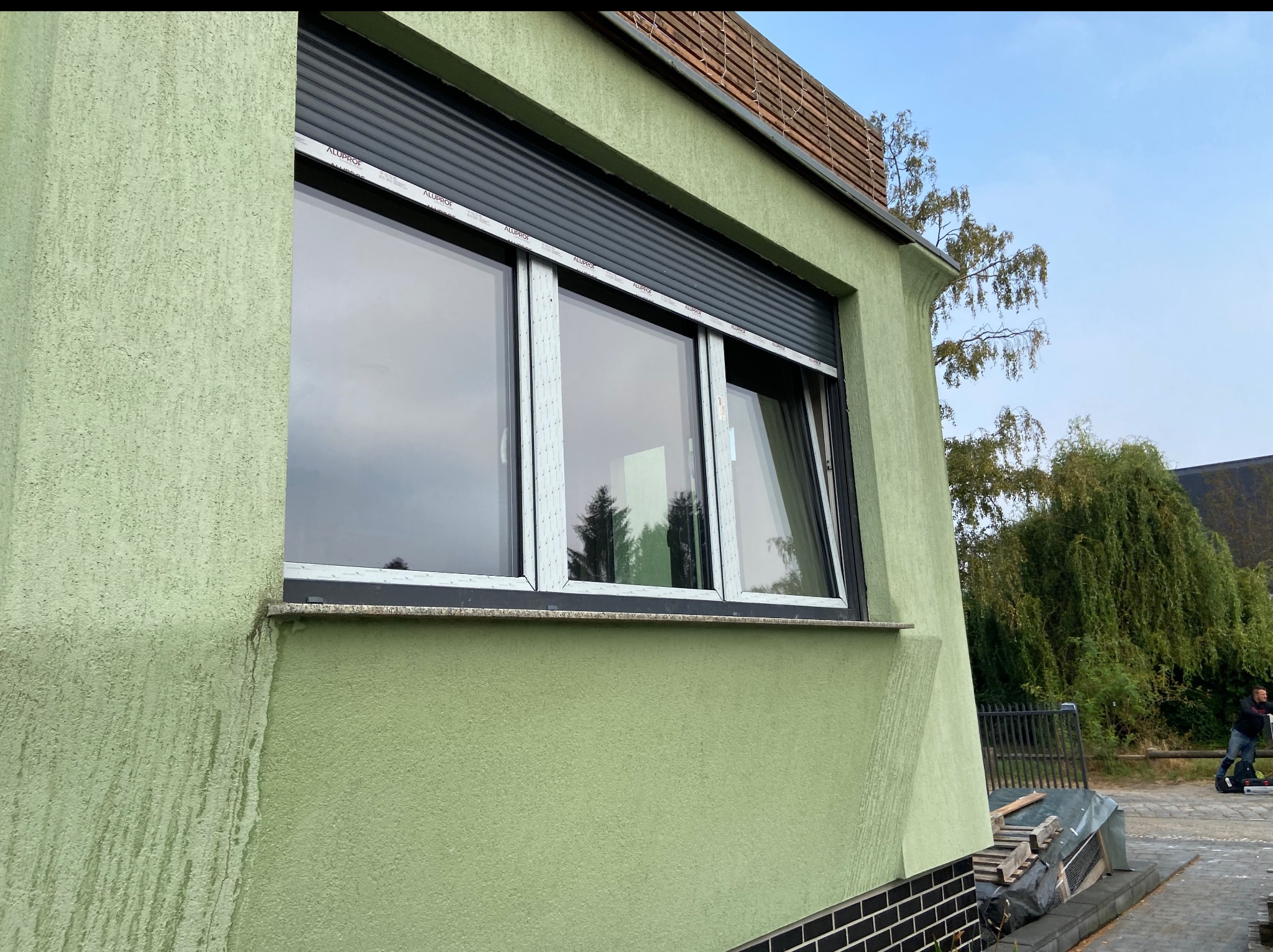 Trzyczęściowe okno PCV z szarą roletą zewnętrzną w zielonym budynku. Jedno skrzydło okna jest otwarte. Widoczne odbicie drzew w szybie.