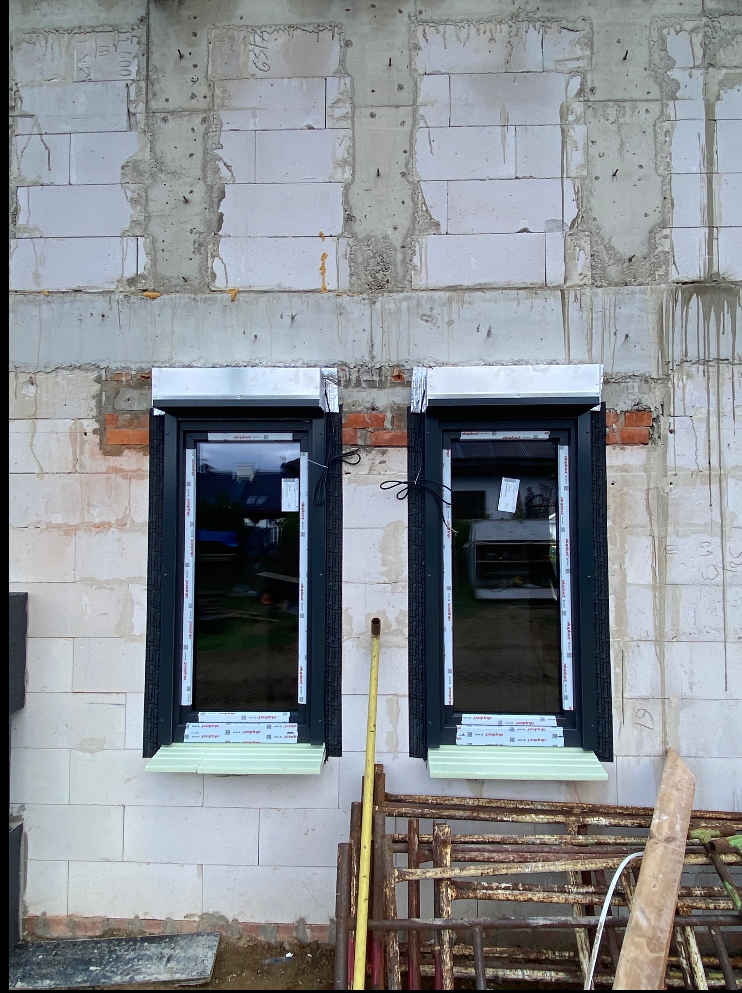 Dwa okna PCV z roletami zewnętrznymi w surowym murze z betonu komórkowego, widoczne elementy montażowe i rusztowanie w tle, okna z szybami refleksyjnymi.