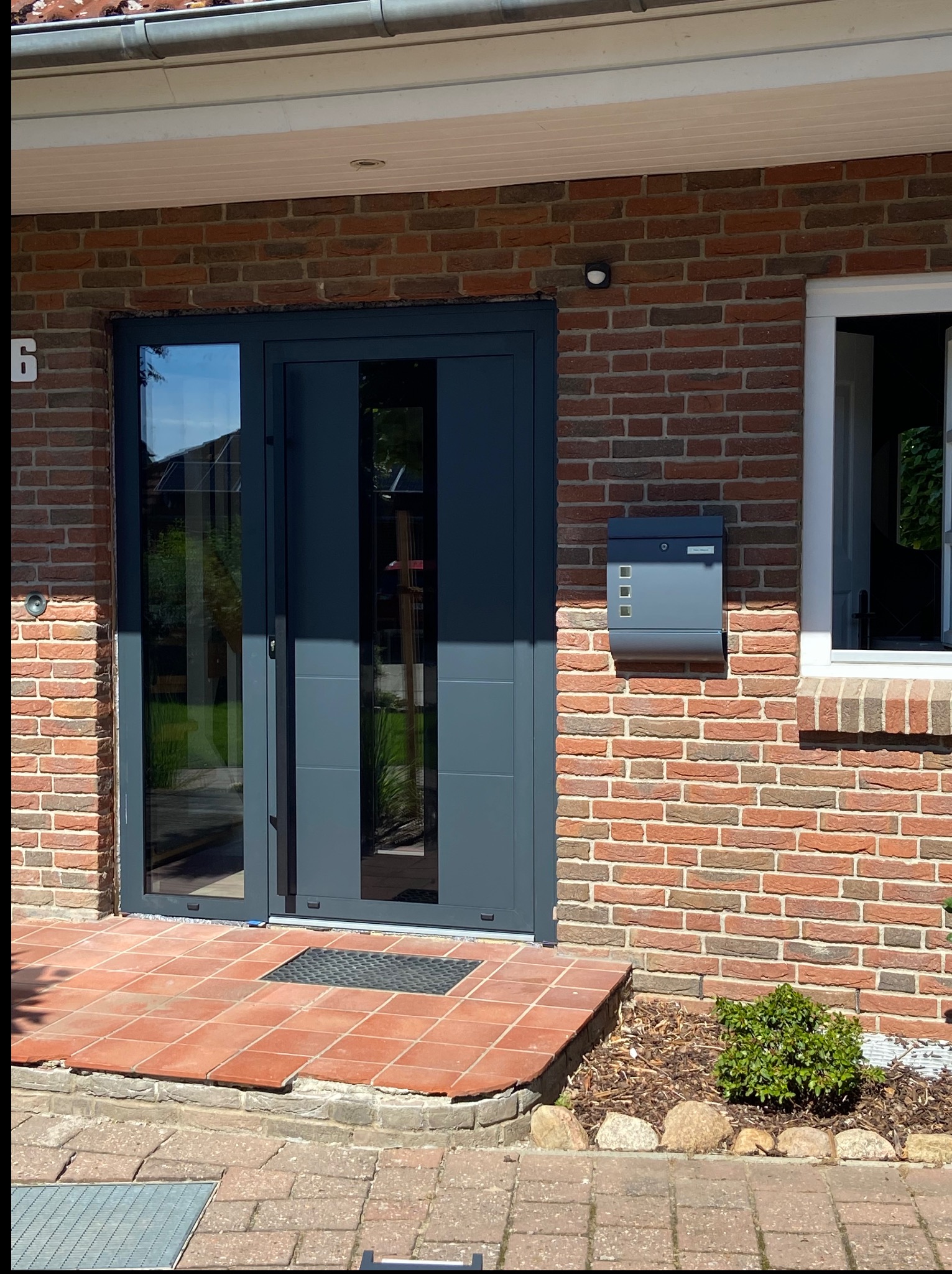 Nowoczesne, grafitowe drzwi wejściowe z przeszkleniami, zamontowane w ceglanym budynku z numerem 6. Obok skrzynka pocztowa w tym samym kolorze. Widoczne płytki wejściowe.