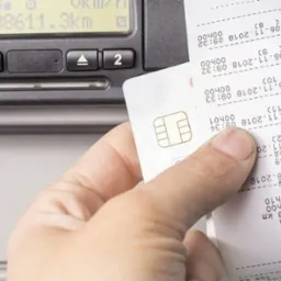 Ręka trzymająca wydruk z tachografu cyfrowego i kartę kierowcy, obok urządzenia tachografu z widocznym wyświetlaczem i przyciskami.