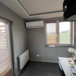 Biały klimatyzator ścienny z wyświetlaczem temperatury, zamontowany nad grzejnikiem w szarym pokoju z oknami z żaluzjami.