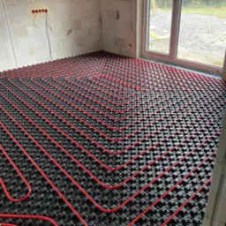 Wnętrze w stanie surowym z ułożonym systemem czerwonych rur na czarnej macie do ogrzewania podłogowego, widok z góry, okno w tle.