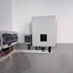 Zamontowany inwerter fotowoltaiczny Fox z podłączonymi przewodami, obok skrzynka rozdzielcza i licznik energii, na tle dwukolorowej ściany (biały i szary).