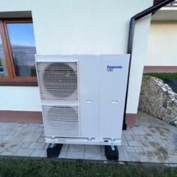 Biała, zewnętrzna jednostka klimatyzacyjna Panasonic, zamontowana na czarnych gumowych podstawach na wybrukowanym tarasie przy ścianie domu z brązowym oknem i rynną.