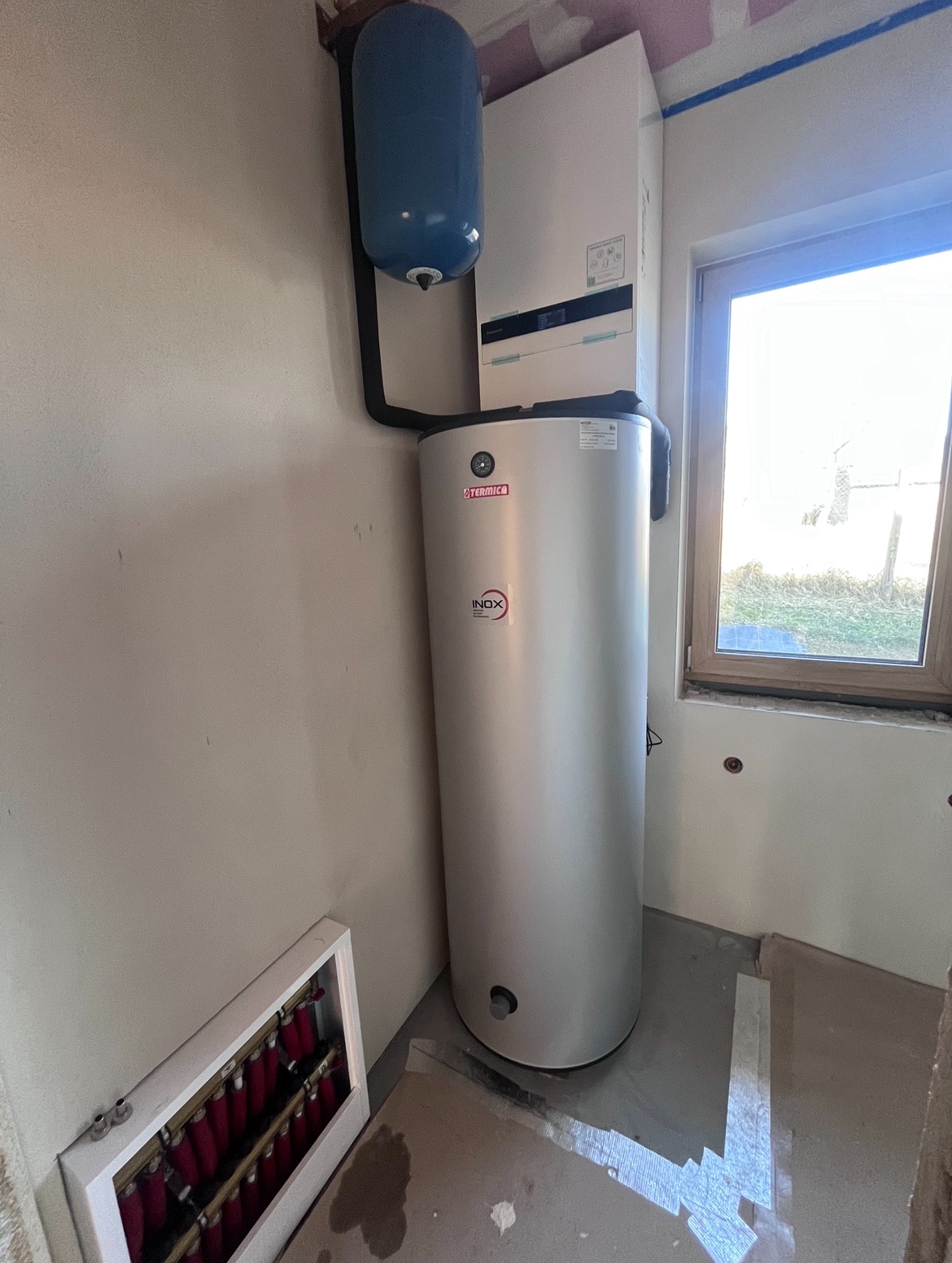 Biały zasobnik ciepłej wody użytkowej marki Termica Inox z niebieskim naczyniem przeponowym, zamontowany obok okna w surowym wnętrzu z widoczną rozdzielnią ogrzewania podłogowego.