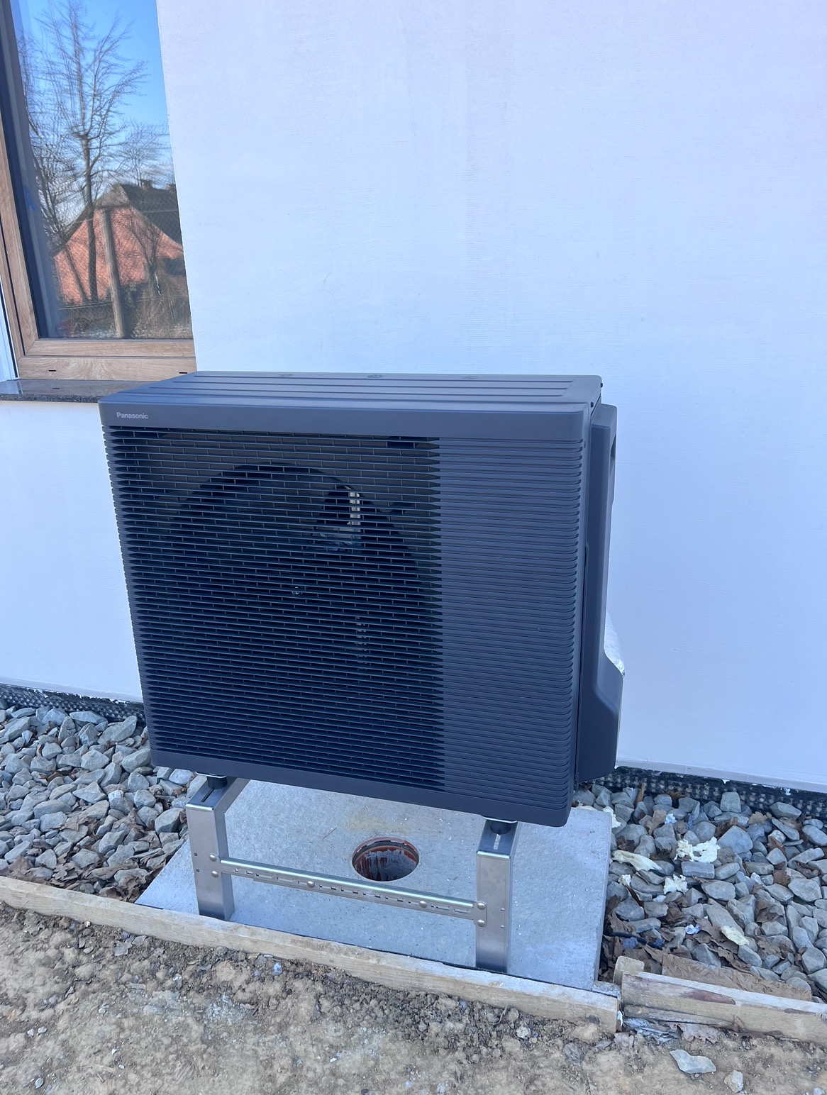 Zewnętrzna jednostka pompy ciepła marki Panasonic, zamontowana na metalowej konstrukcji na betonowej podstawie, widoczna na tle białej ściany budynku i szarego żwiru.