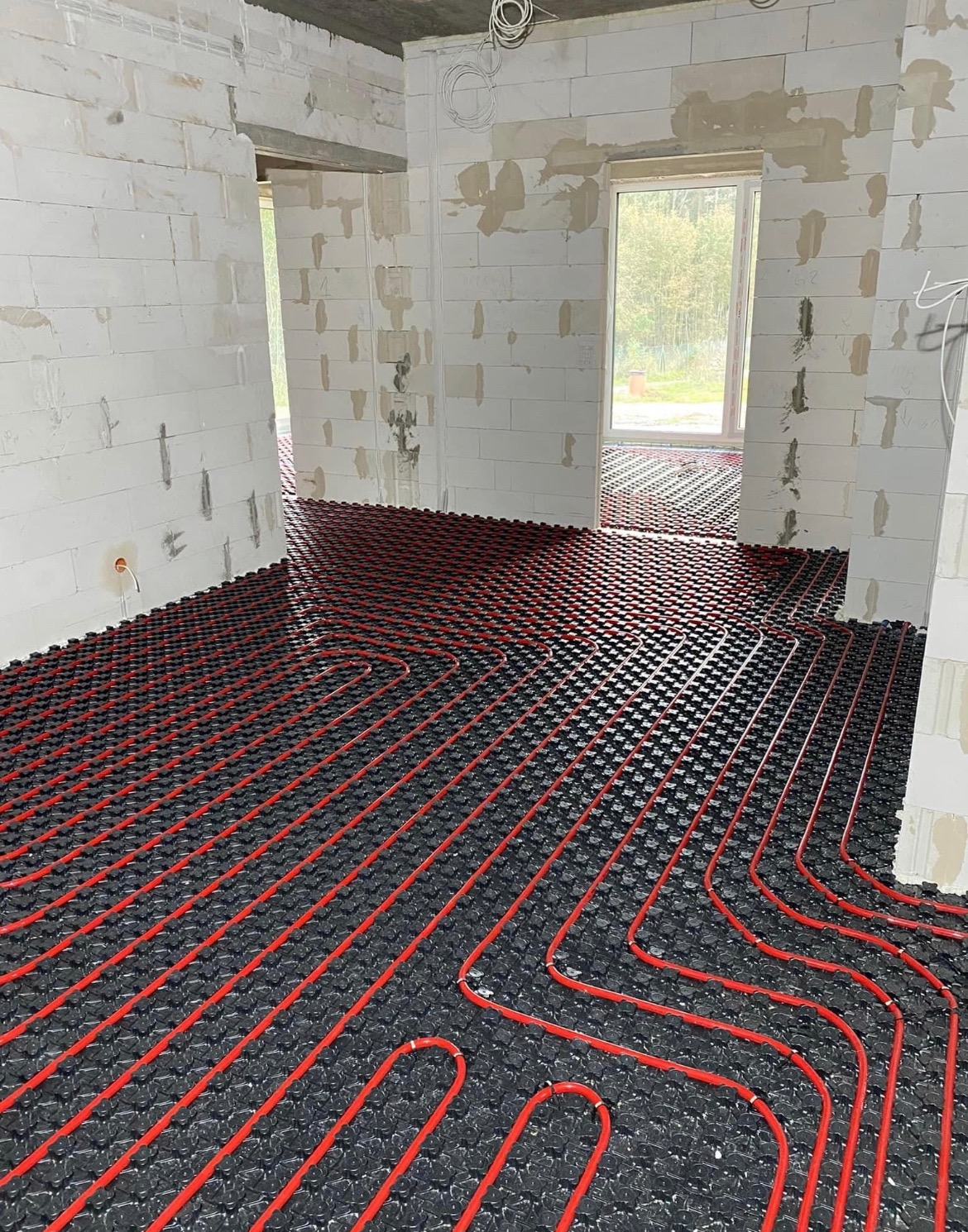 Czerwone rurki ogrzewania podłogowego ułożone na czarnej izolacji w surowym wnętrzu budynku z białych bloczków, widoczne okno w tle.