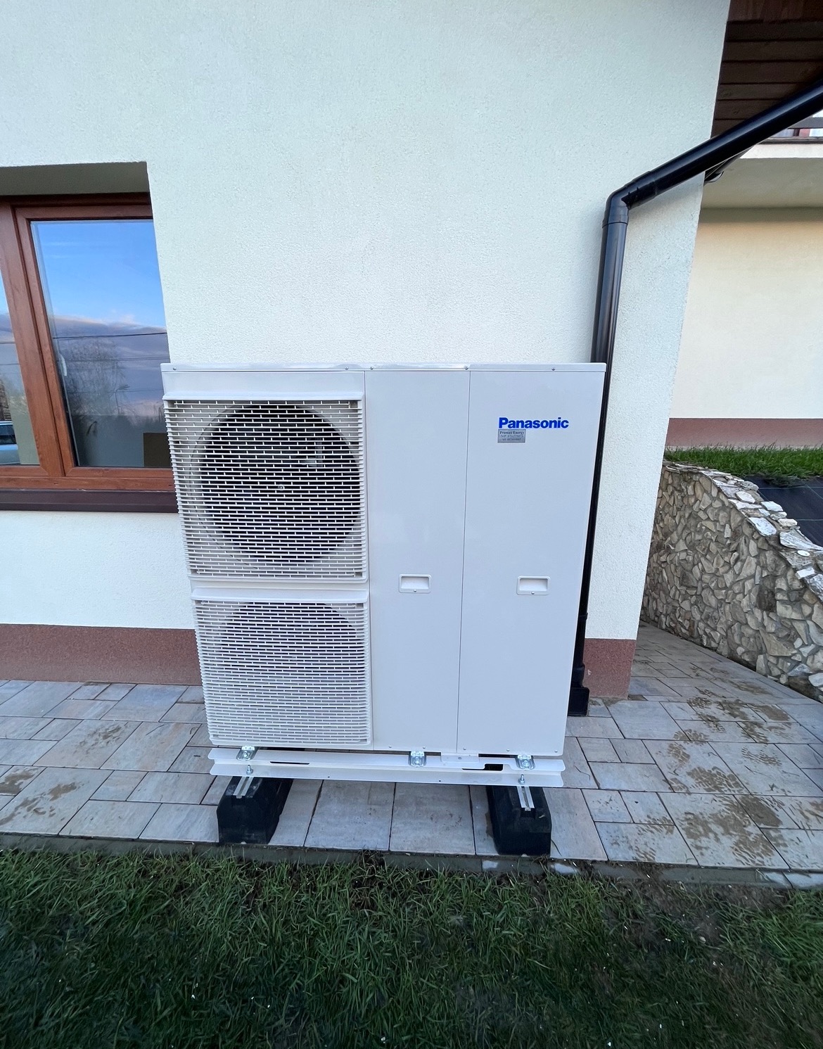 Biała, zewnętrzna jednostka klimatyzacyjna Panasonic, zamontowana na czarnych gumowych podstawach na wybrukowanym tarasie przy ścianie domu z brązowym oknem i rynną.