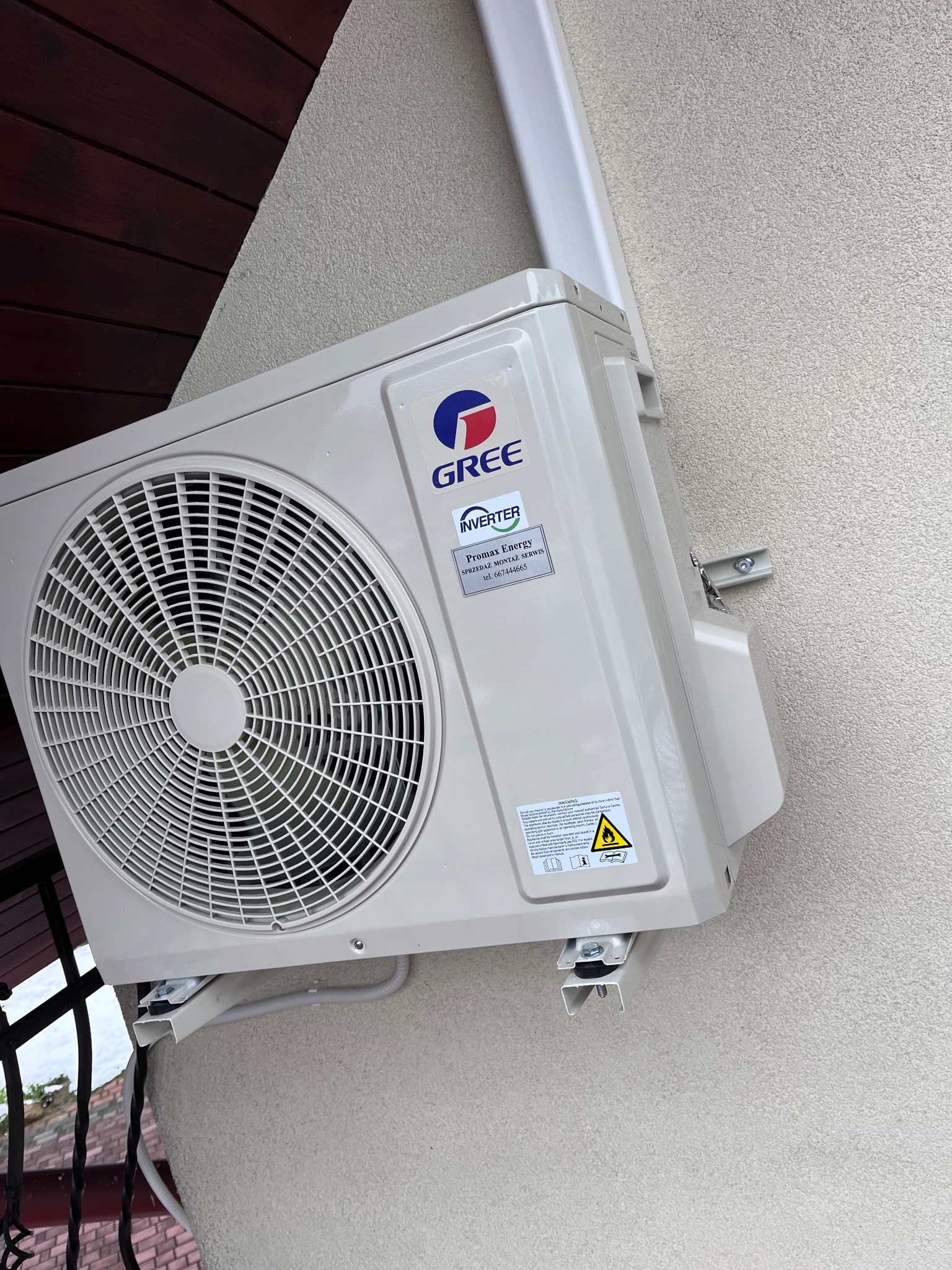 Zewnętrzna jednostka klimatyzacji marki Gree zamontowana na ścianie budynku z widocznym fragmentem orynnowania i podbitki dachowej.
