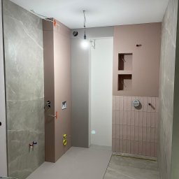 Usługi Ogólnobudowlane Andrii Fryz - Wnętrze łazienki w trakcie remontu: szare płytki imitujące marmur na podłodze i ścianie, różowe ściany z wnękami, pionowe różowe kafelki przy armaturze, widoczne rury i instalacje, surowy stan...