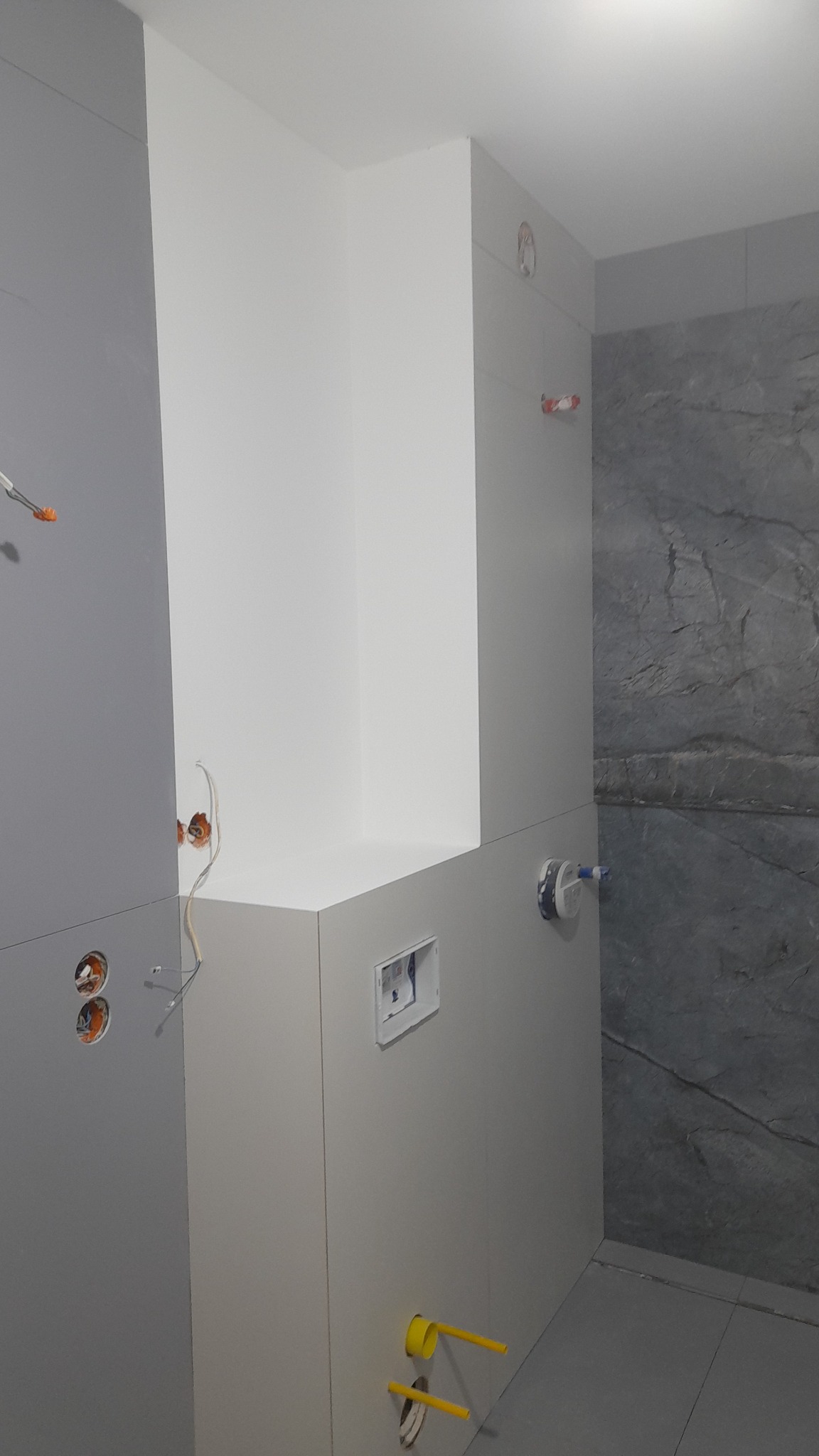 Szare i białe płytki w łazience w trakcie wykańczania, widoczne wystające kable elektryczne i rury kanalizacyjne.