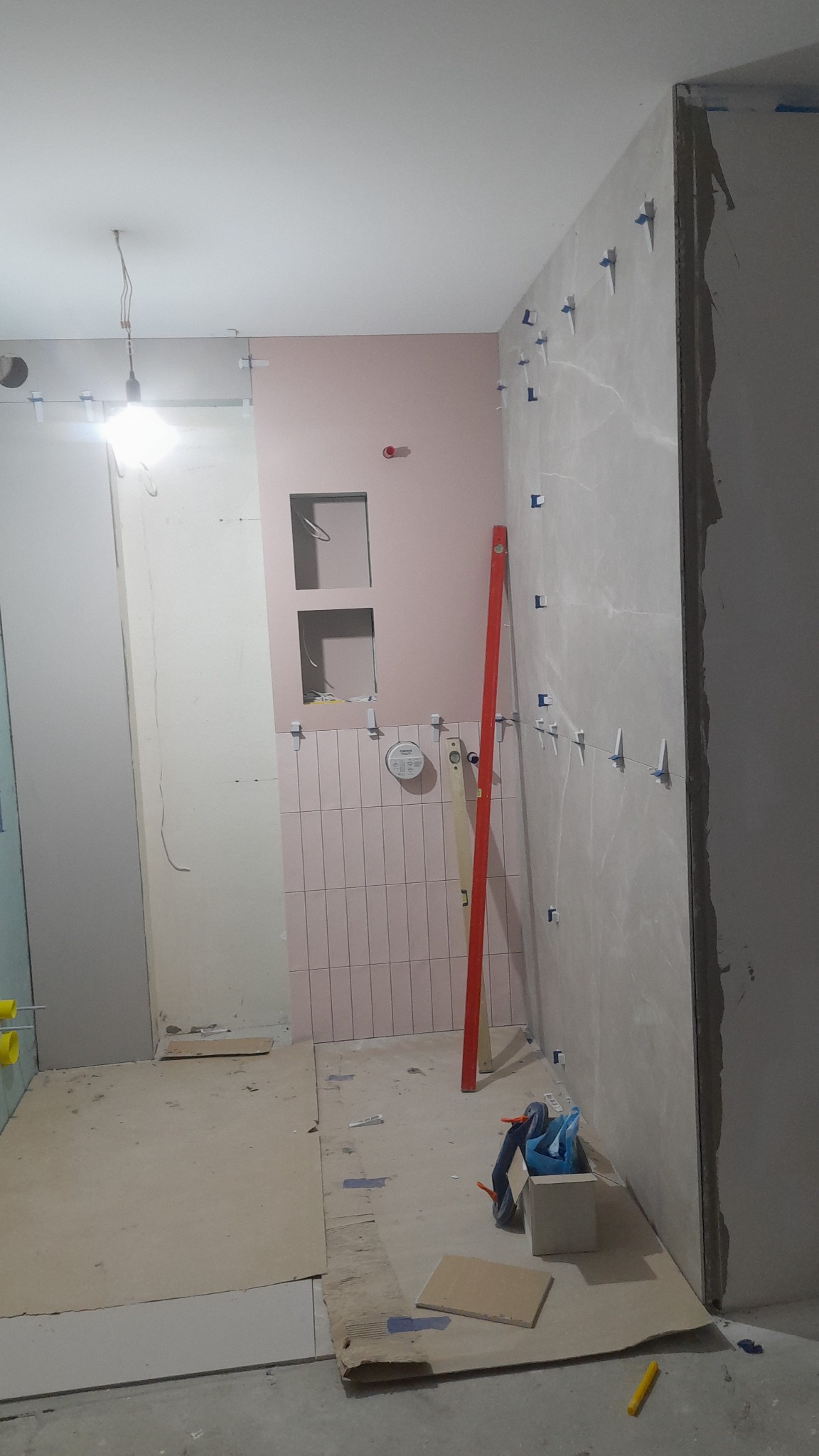 Łazienka w trakcie remontu: różowe płytki ułożone pionowo na ścianie z wnękami, obok ściana z płyt kartonowo-gipsowych z niebieskimi krzyżykami dystansowymi, poziomica oparta o ścianę, podłoga...