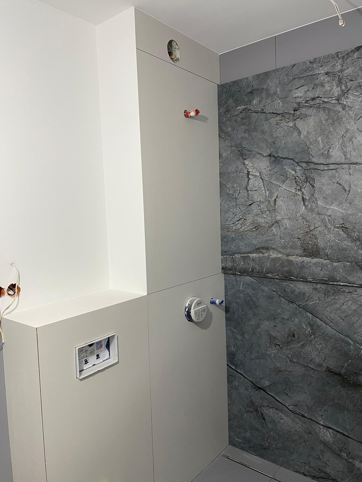 Szara marmurowa ściana prysznicowa sąsiaduje z białą zabudową WC z widocznymi instalacjami i otworem na przycisk spłukiwania.