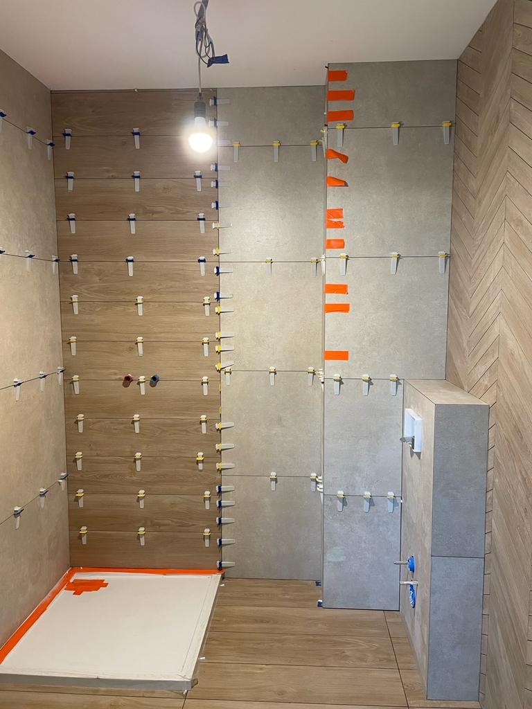 Układanie płytek imitujących drewno i szarych płytek w łazience, widoczne poziomowanie z użyciem klipsów i klinów, brodzik zabezpieczony taśmą malarską.