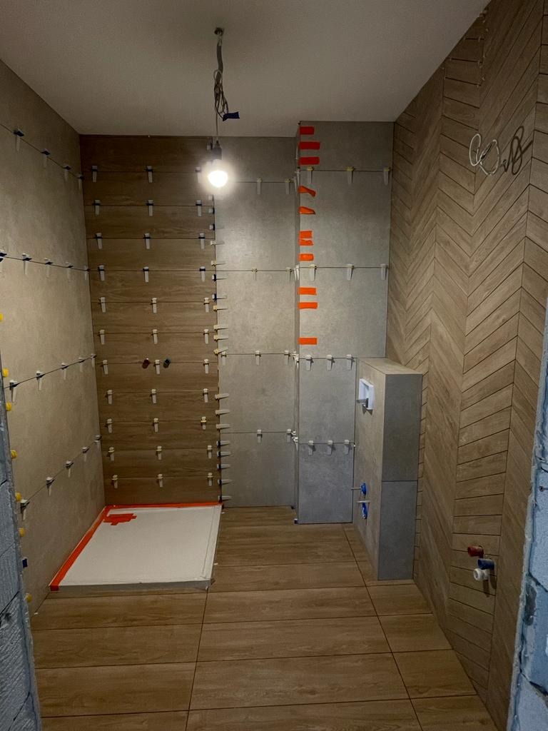 Łazienka w trakcie wykańczania: brązowe płytki imitujące drewno na podłodze i ścianie ułożone we wzór jodełki, szare płytki na pozostałych ścianach, widoczne krzyżyki dystansowe i poziomice...