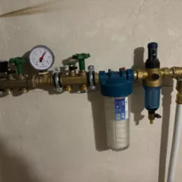Złożony system filtracji wody z manometrem i zaworami, zamontowany na surowej, pomalowanej ścianie. Widoczne plastikowe rury i przezroczyste obudowy filtrów z wkładami.