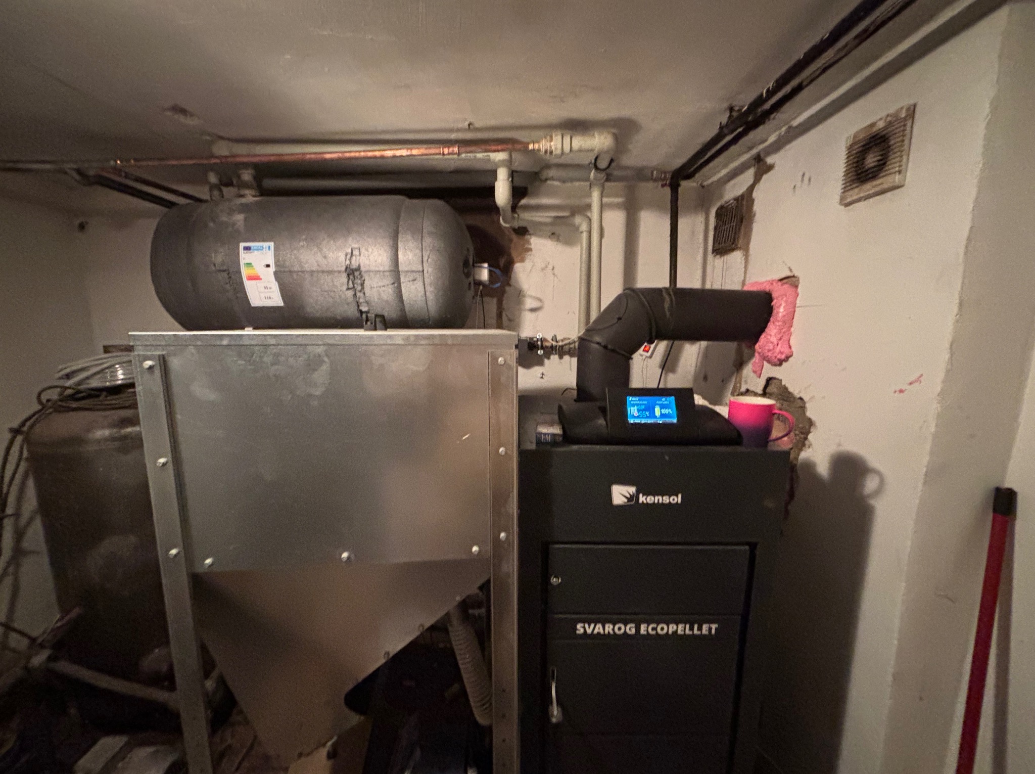 Kocioł na pellet Svarog Eco Pellet z podajnikiem, zbiornikiem na wodę i rurami w piwnicy, widoczny kubek na górze kotła.