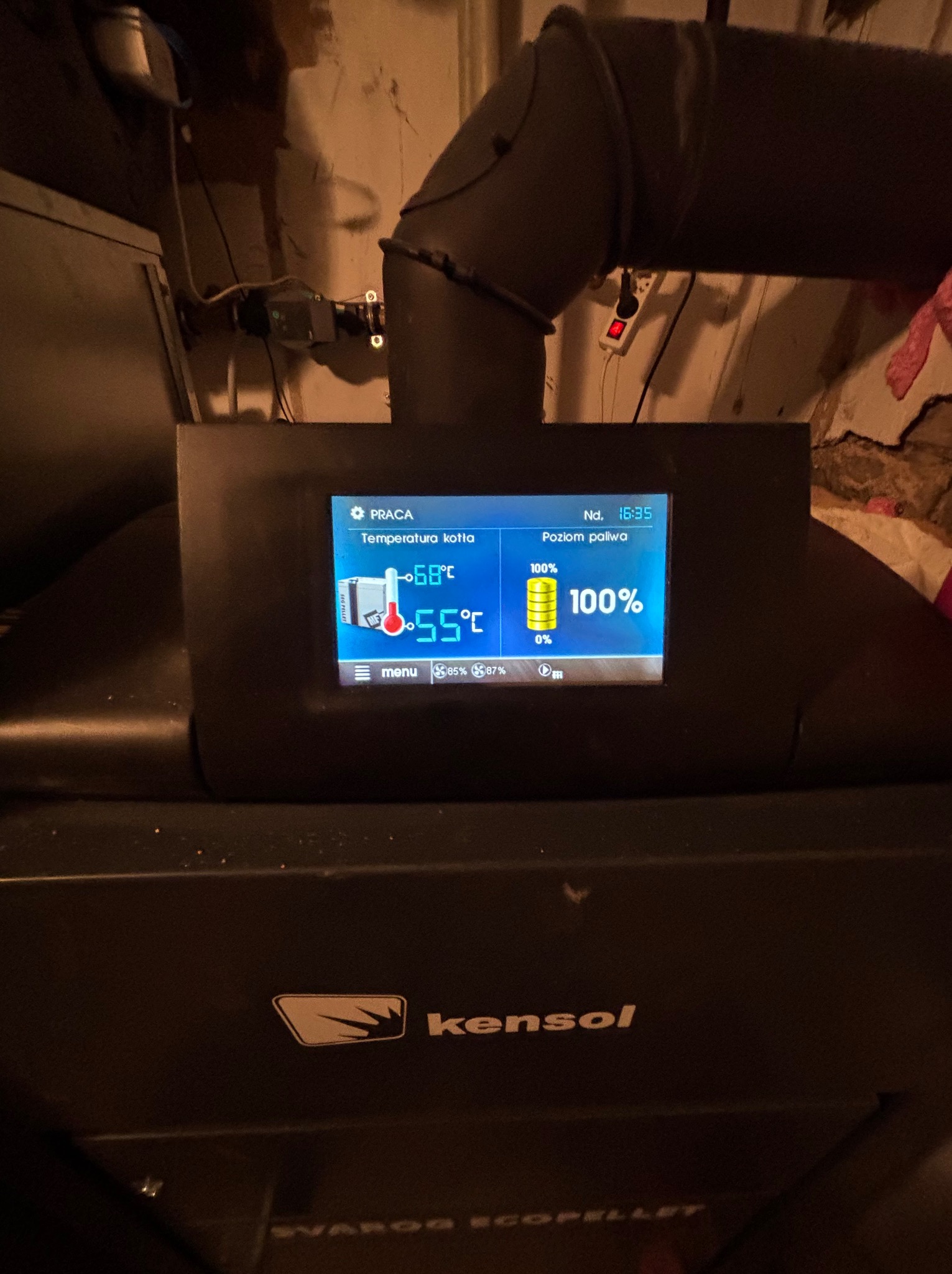 Panel sterowania pieca na pellet Kensol Svarog Ecopellet z widoczną temperaturą kotła 55 stopni Celsjusza i poziomem paliwa 100%, rura odprowadzająca spaliny w tle.