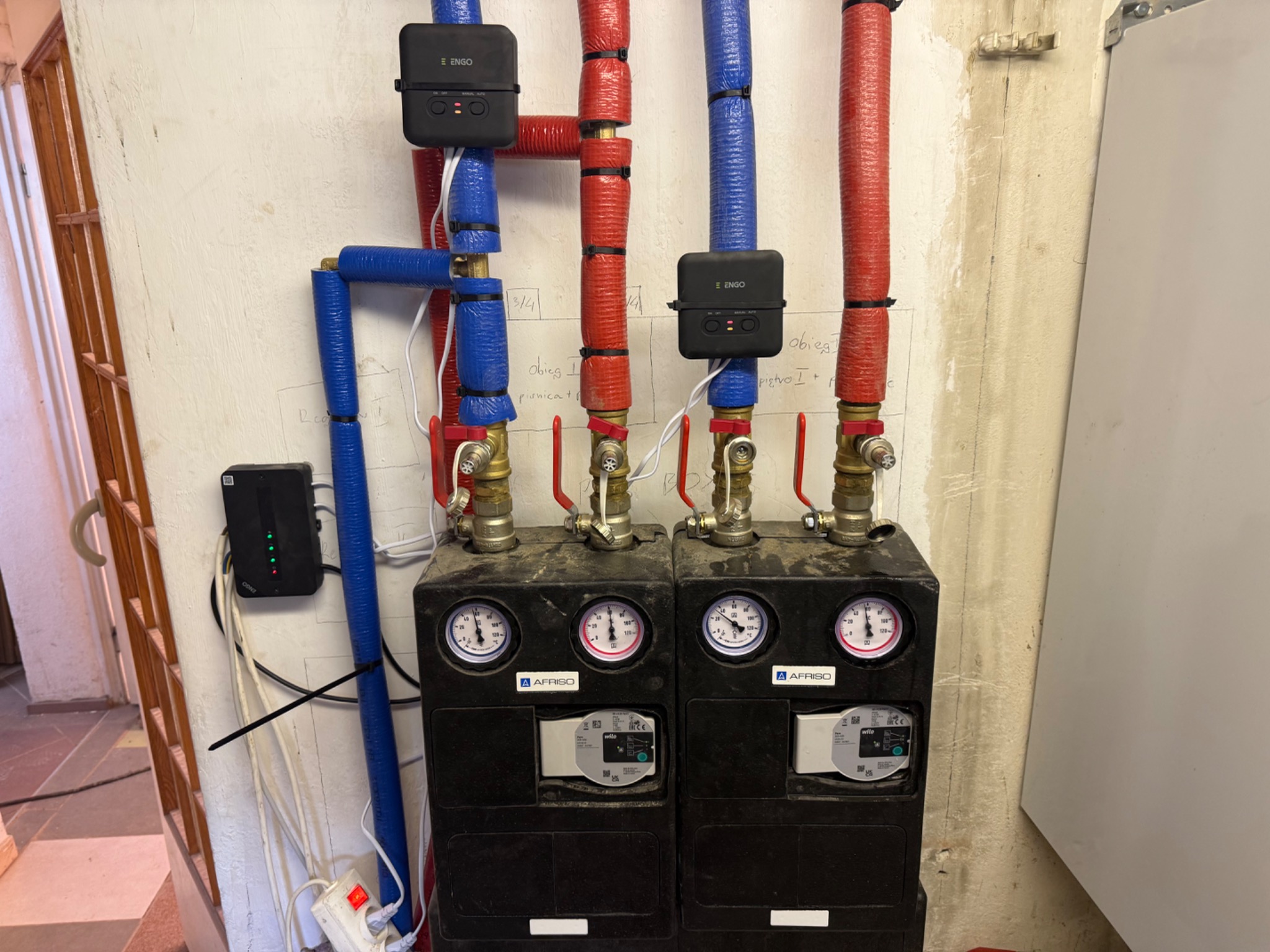Dwa czarne, kompaktowe moduły AFRISO z manometrami, połączone rurami w izolacji (czerwona i niebieska), sterowane urządzeniami ENGO. Instalacja w pomieszczeniu.