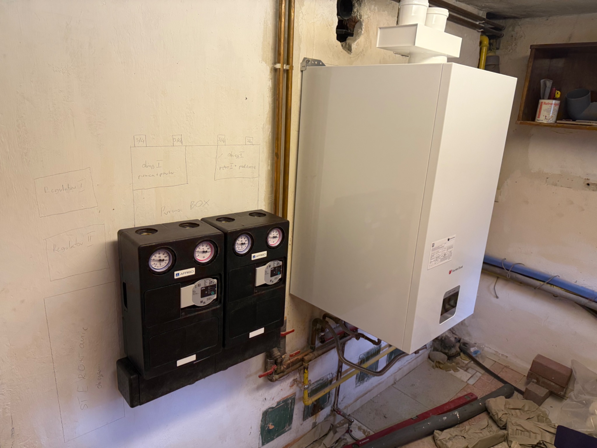 Nowoczesny piec gazowy Saunier Duval z czarnym sterownikiem Purmo BOX na ścianie z instalacją. Widoczne rury miedziane i schemat instalacji narysowany na ścianie.