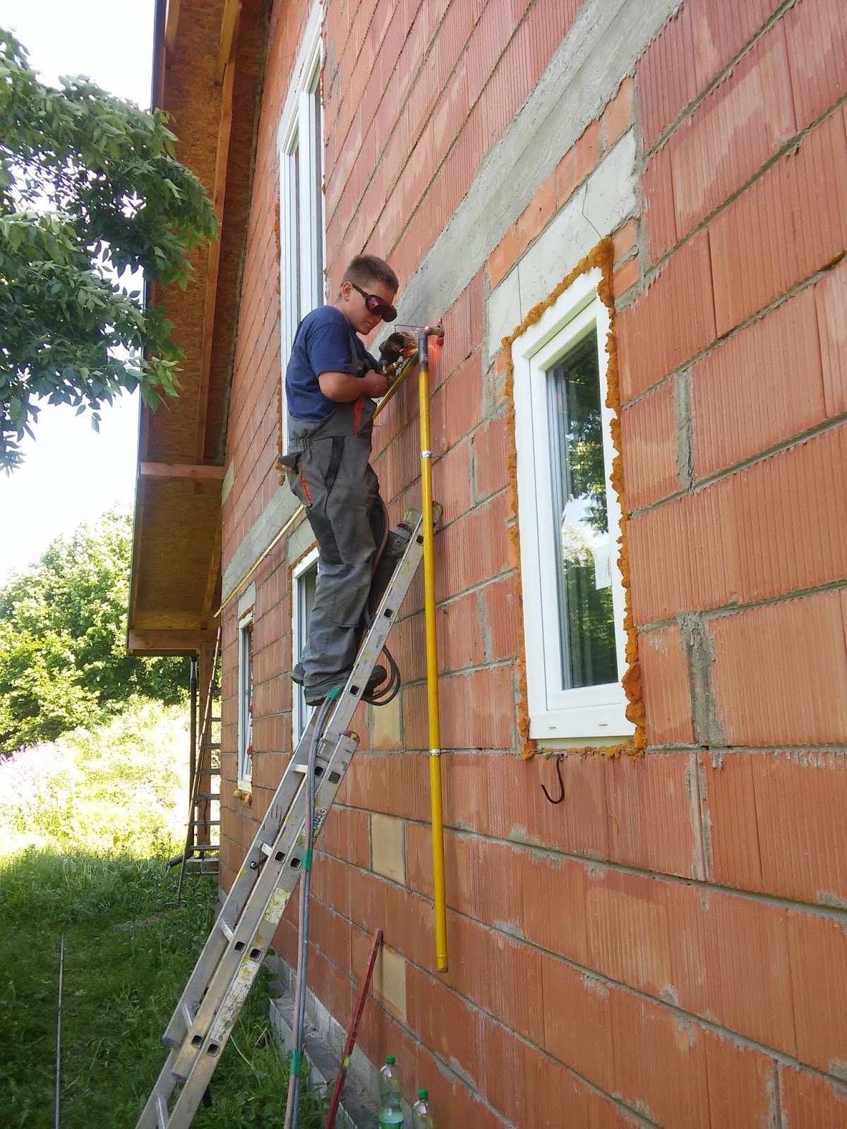 Pracownik na drabinie montuje instalację gazową przy oknie w budynku z czerwonej cegły. Widoczne narzędzia i elementy instalacji.