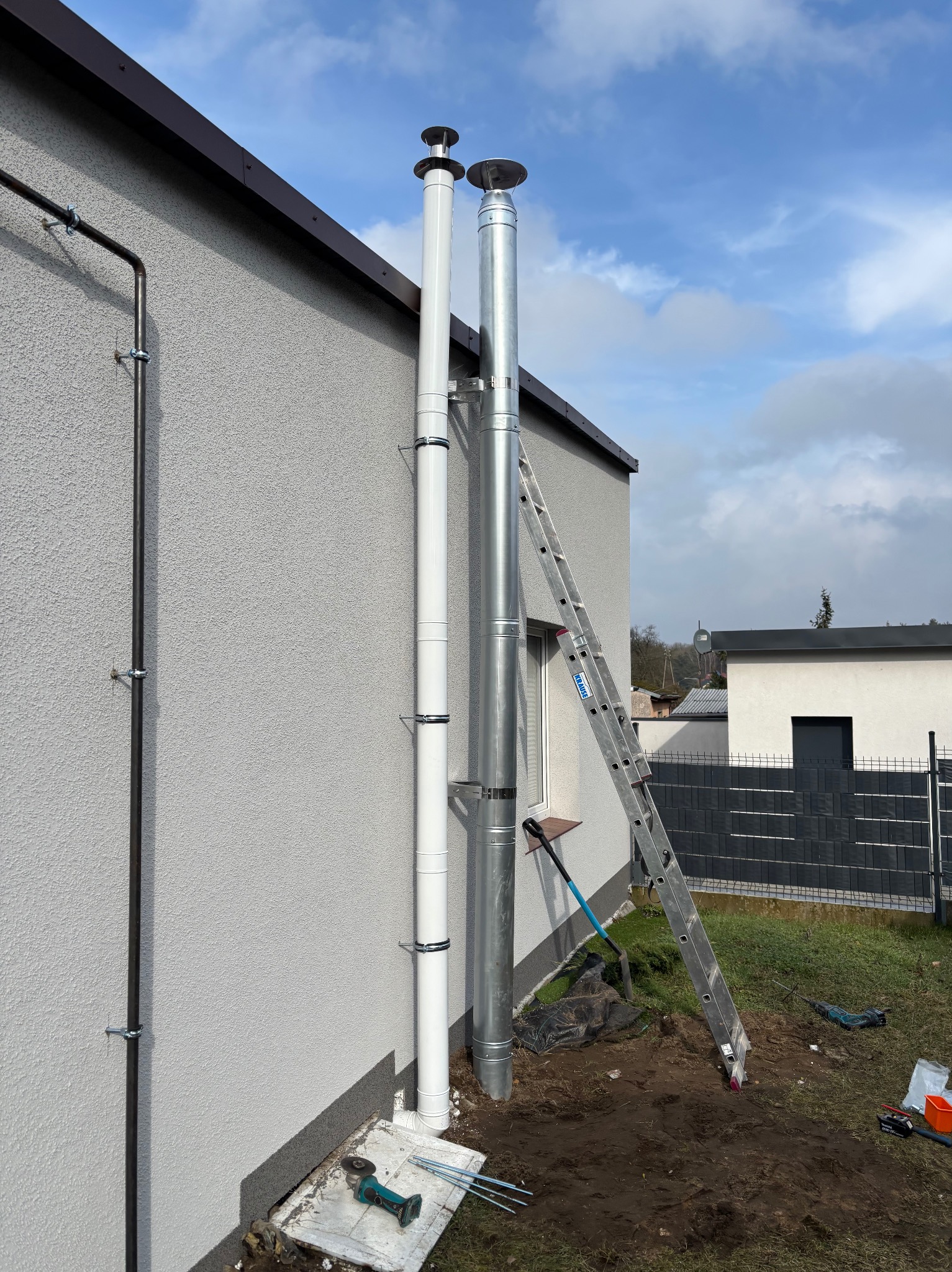 Dwa kominy zewnętrzne (biały i metalowy) zamontowane przy ścianie budynku. Aluminiowa drabina oparta o ścianę. Widoczne narzędzia i elementy instalacji.