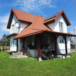 Dwupiętrowy dom z czerwoną dachówką i panelami słonecznymi na dachu, widok z trawnika w słoneczny dzień.