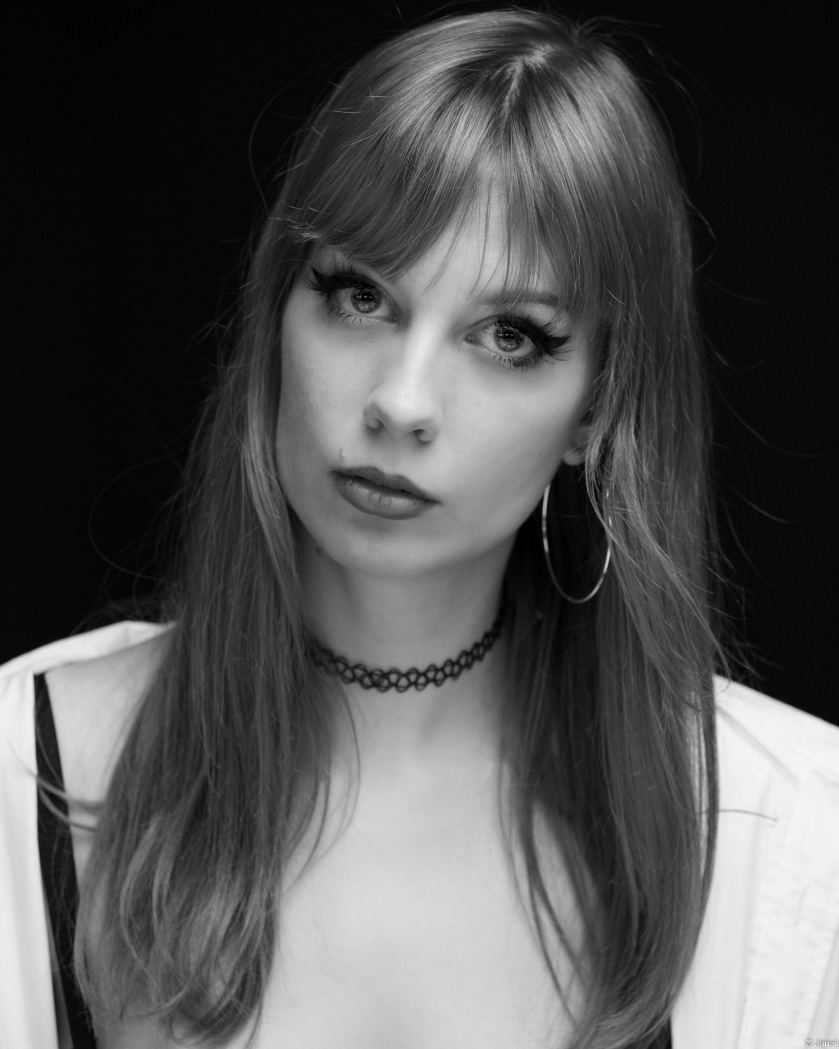 Portret czarno-biały młodej kobiety z długimi włosami, chokerem i kolczykami kołami, na czarnym tle.