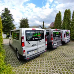 Rząd srebrnych i szarych busów marki Renault Trafic zaparkowanych na betonowej kostce z logo firmy przewozowej i numerem telefonu na tylnej szybie, z napisem 'Niemcy - Belgia - Holandia - Szwajcaria'.