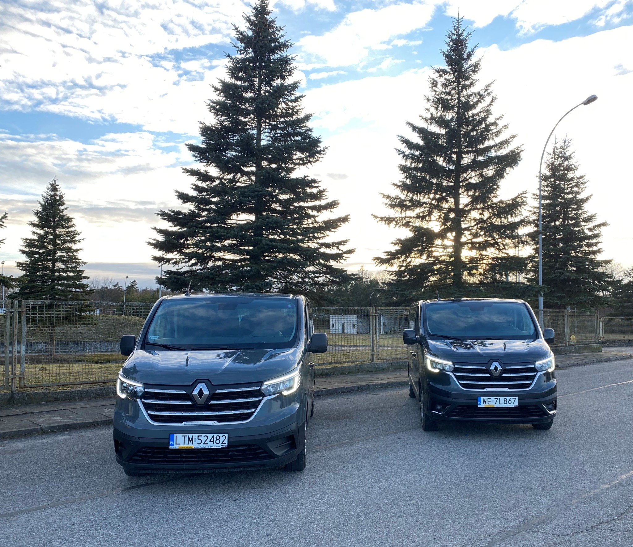 Dwa ciemne busy Renault Trafic zaparkowane na asfalcie, w tle wysokie sosny i pochmurne niebo.