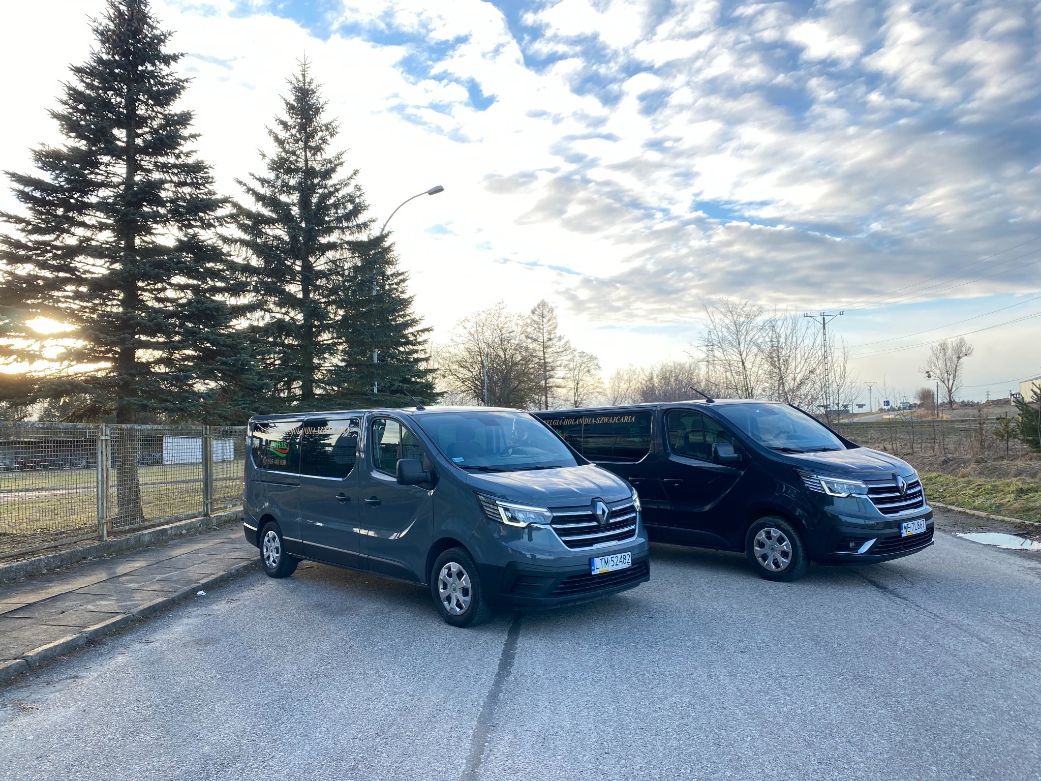 Dwa busy Renault Trafic, szary i czarny, zaparkowane obok siebie na parkingu, w tle drzewa iglaste i pochmurne niebo.